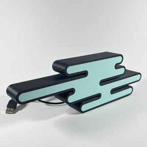 Può includere: Una stazione di ricarica USB nera e blu chiaro a forma di nuvola stilizzata. La stazione di ricarica ha un cavo USB.