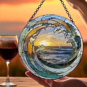 May include: A round stained glass art piece depicting a wave with a sunset scene. The artwork features shades of blue, green, and yellow, with a black chain for hanging. A glass of red wine sits on a wooden surface.