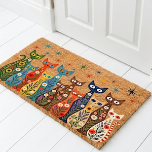 May include: A rectangular tan coir doormat featuring a row of eight stylized cats in various colors including green, blue, orange, yellow, brown, and white. Each cat is decorated with floral patterns and surrounded by small colorful starburst icons.