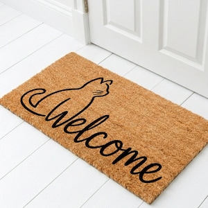 May include: A rectangular tan coir doormat featuring a black line art illustration of a sitting cat and the word Welcome written in a cursive script font. The mat is positioned on a white floor in front of a white door with a bouquet of white roses.