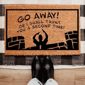 May include: A rectangular doormat made of natural coir fiber. The mat features the text "GO AWAY! OR I SHALL TAUNT YOU A SECOND TIME!" in black, along with a silhouette of a person and brick walls. The mat is placed on a black and white checkered rug.