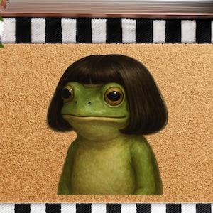 May include: A doormat featuring a green frog with large eyes and a dark brown bob haircut. The frog is centered on a tan background, with a black and white striped border. The doormat is a decorative item.