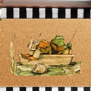 May include: A rectangular doormat with a light brown background features an illustration of two frogs in a boat. One frog reads a book, while the other fishes. The design includes a lily pad and a black and white striped border.