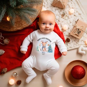 Puede incluir: Un body blanco de bebé con mangas largas con un gráfico de ciervo navideño y el texto "Baby's First Christmas", el nombre "James" y el año "2024". El body está sobre una manta roja.