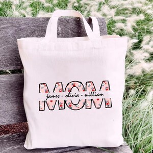 Puede incluir: Bolsa de tela blanca con la palabra "MOM" en letras negras delineadas. Las letras están rellenas con un patrón floral rosa. Debajo de las letras hay una pancarta rosa con el texto "james - olivia - william" en negro.
