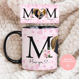 Puede incluir: Una taza rosa y negra con una gran letra "M" decorada con flores rosas y un marco de fotos en forma de corazón. La taza tiene un asa negra y el texto "Te amo" está impreso en la taza. El fondo es un patrón rosa de corazones.