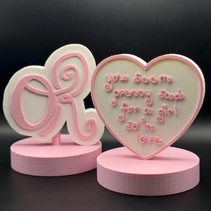 May include: Two pink and white decorative items. One is a cloud-shaped design with the letters "OR" in pink. The other is a heart shape with the text "You seem pretty good for a girl so in love" in pink.