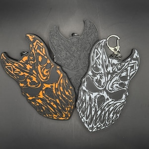 Könnte beinhalten: Drei Schlüsselanhänger mit einem wolfsähnlichen Schädeldesign. Einer ist orange und schwarz, ein weiterer grau und der dritte schwarz. Jeder Schlüsselanhänger hat einen Metallring zum Befestigen an Schlüsseln oder einer Tasche. Die Schlüsselanhänger sind aus Gummi.