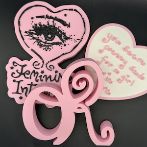 Può includere: Oggetti decorativi rosa con forme di cuore e testo. Un cuore ha un disegno di occhio, un altro dice "You seem pretty good for a girl", e un terzo ha le parole "Feminine Intellect". Anche la lettera "R" è rosa.