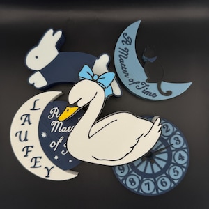 Könnte beinhalten: Dekorative Holzausschnitte in Weiß und Marineblau. Enthält einen Schwan mit blauer Schleife, ein Kaninchen, eine Katze auf einem Mond, eine Uhr und einen Mond mit dem Text "A Matter of Time" und "LAUFEY".