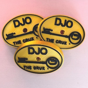 以下が含まれることがあります： 黒い鍵のデザインと「DJO THE CRUX.」という文字が書かれた、黄色い楕円形のキーホルダーが3つあります