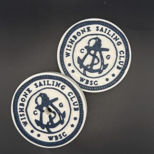Könnte beinhalten: Zwei runde Untersetzer mit einem Ankerdesign in Marineblau und dem Text "Wishbone Sailing Club". Die Untersetzer haben einen weißen Hintergrund mit einem marineblauen Rand und den Buchstaben "WBSC".