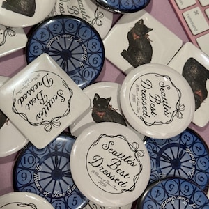 Könnte beinhalten: Eine Sammlung runder und quadratischer Knöpfe und Anstecknadeln. Einige zeigen eine schwarze Katze mit roter Fliege, andere ein Zifferblatt-Design mit Zahlen, und einige haben den Text "Seattle's Best Dressed".