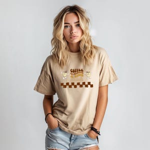 Puede incluir: Camiseta color beige con la frase "Coffee is Always a Good Idea" en una fuente retro. El diseño incluye un patrón de tablero de ajedrez marrón y personajes de tazas de café. La modelo lleva vaqueros azules.