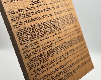 以下が含まれることがあります： 黒いインクで刻まれた米国憲法のテキストが書かれた木製の銘板。銘板は黒い金属製のスタンドの上に置かれています。