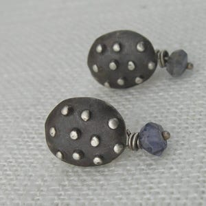 Peut inclure: Une paire de boucles d'oreilles en argent avec une base ronde en métal gris texturé et un petit pendentif en pierre précieuse violette foncé.
