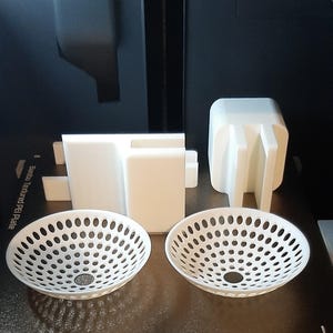 Pode incluir: Uma coleção de objetos brancos impressos em 3D, incluindo duas tigelas perfuradas, uma peça retangular e uma estrutura quadrada com suportes verticais. Os itens estão dispostos em uma superfície escura, provavelmente uma base de impressora.