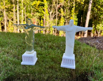 May include: Two Christ the Redeemer statues standing side by side. One statue is made of clear, transparent material, while the other is made of solid white material. Both figures feature outstretched arms and are mounted on white rectangular bases.