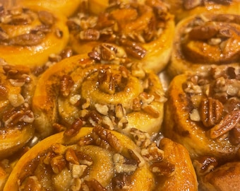 May include: A batch of golden-brown cinnamon rolls arranged in a foil pan, each spiral pastry topped with a generous amount of chopped pecans and a glossy, sticky glaze that coats the dough and fills the crevices between the individual rolls.
