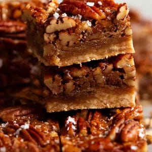 Könnte beinhalten: Nahaufnahme von gestapelten Pekannuss-Kuchenriegeln. Die Riegel haben einen goldbraunen Boden, eine Schicht Pekannussfüllung und sind mit ganzen Pekannüssen und grobem Salz belegt. Ein süßer Leckerbissen.