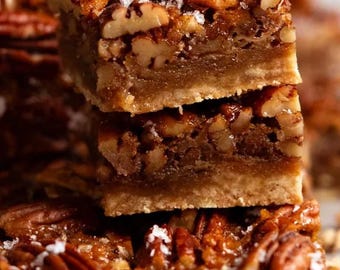 May include: Close-up of pecan pie bars stacked on top of each other. The bars have a golden-brown crust, a layer of pecan filling, and are topped with whole pecans and coarse salt. The dessert is a sweet treat.
