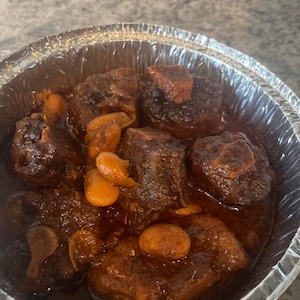 Puede incluir: Primer plano de un recipiente de aluminio lleno de rabo de buey estofado. El rabo de buey es de color marrón oscuro, brillante con salsa y entremezclado con dientes de ajo enteros. El plato parece sabroso y listo para comer.