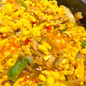 Puede incluir: Primer plano de una sartén de ackee y bacalao salado jamaicano, un plato tradicional caribeño. El plato está hecho con fruta de ackee, bacalao salado, cebollas, pimientos y especias. El ackee es de color amarillo brillante y el bacalao salado es blanco y escamoso. El plato está hirviendo a fuego lento en una salsa a base de tomate.