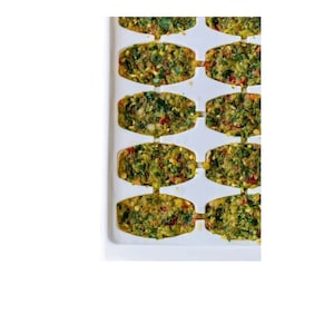 May include: A white tray filled with rectangular portions of a green, textured food mixture. The food appears to contain herbs, spices, and possibly vegetables. The portions are separated by narrow channels within the tray.