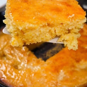 May include: A square slice of golden-brown cornbread is lifted from a cast iron skillet with a spatula. The cornbread has a slightly glossy, caramelised top and a crumbly texture. The remaining cornbread in the skillet is also golden brown.