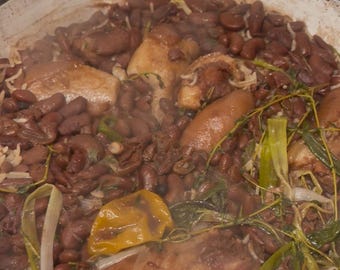 May include: A pot of Jamaican-style red beans and rice, with visible ingredients including red kidney beans, chicken, rice, and vegetables. The dish is steaming, indicating it is freshly cooked and ready to serve. The pot is white with a dark rim.