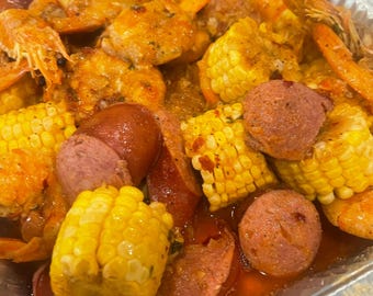 May include: A foil container filled with a seafood boil. The image shows cooked prawns, sweetcorn on the cob, and sliced sausage. The prawns are orange, the sweetcorn is yellow, and the sausage is pink. The food is coated in a sauce.
