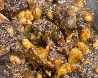 May include: A hearty stew consisting of dark, braised meat chunks with visible bone marrow and light-colored, oval-shaped beans coated in a thick, savory, dark brown sauce. The dish is presented in a clear plastic container with visible herbs.