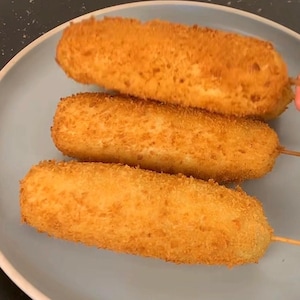 Puede incluir: Tres perros de maíz dorados sobre un plato azul claro. Los perros de maíz están fritos para obtener una textura crujiente y están ensartados con palitos de madera. Un aperitivo popular.