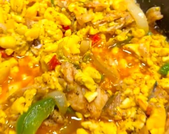 May include: A close-up of a pan of Jamaican ackee and saltfish, a traditional Caribbean dish. The dish is made with ackee fruit, salt cod, onions, peppers, and spices. The ackee is bright yellow, and the salt cod is white and flaky. The dish is simmering in a tomato-based sauce.