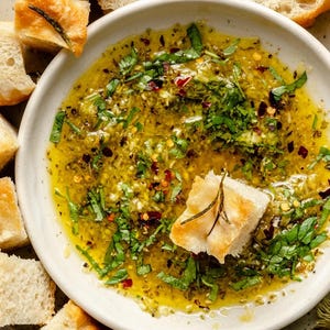 May include: A white ceramic bowl filled with olive oil, herbs, and spices, surrounded by toasted bread cubes. The oil is a vibrant yellow-green, with visible herbs and red pepper flakes. A piece of bread sits in the oil.