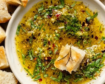 May include: A white ceramic bowl filled with olive oil, herbs, and spices, surrounded by toasted bread cubes. The oil is a vibrant yellow-green, with visible herbs and red pepper flakes. A piece of bread sits in the oil.
