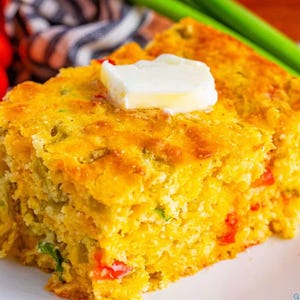 Puede incluir: Una rebanada cuadrada de pan de maíz amarillo dorado cubierta con una porción rectangular de mantequilla blanca. El pan de maíz tiene una superficie texturizada con motas verdes de hierbas y trozos de pimiento rojo en la miga.