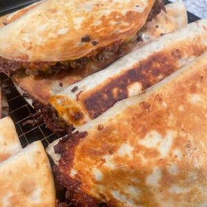 Può includere: Primo piano di diverse quesadillas dorate, alcune piegate e altre piatte, ripiene di un ripieno saporito. Le tortillas hanno una consistenza croccante e leggermente carbonizzata, che suggerisce che sono state cotte su una piastra. Le quesadillas sono disposte su una griglia nera.