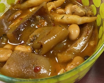 May include: A close-up shot of a bowl filled with a stew-like dish. The stew contains large, irregularly shaped pieces of what appears to be meat, along with white beans and other ingredients in a rich, brown broth. The bowl is clear with a yellow and green pattern.
