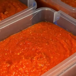 May include: Close-up of several clear plastic containers filled with a vibrant, thick, red sauce. The sauce appears to be freshly made, with a slightly textured surface. The containers are arranged closely together, suggesting a batch of homemade food.
