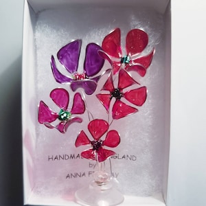 May include: A glass flower bouquet with pink and purple flowers in a clear glass vase. The flowers are handmade by Anna F. and the vase is on a white surface. The text "Handmade by Anna F. England" is visible on the white surface.