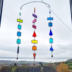 May include: A rainbow-coloured stained glass mobile with geometric shapes hanging from a metal frame. The mobile features a variety of shapes, including squares, rectangles, and triangles, and is suspended from a silver metal frame. The mobile is hanging in front of a window with a view of a cloudy sky and a distant landscape.