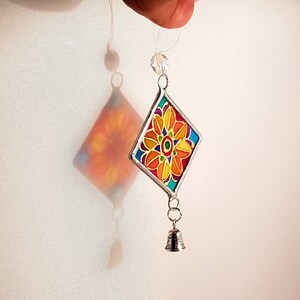 May include: A colorful stained glass diamond-shaped ornament with a floral design in shades of orange, yellow, red, green, and blue. The ornament is suspended from a silver chain with a small silver bell at the bottom.