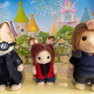 May include: Three flocked animal figurines standing in a row. The left figure is a cat wearing black sunglasses and a black coat. The center figure is a small rabbit wearing a red vest. The right figure is a bear with brown hair wearing a dark blue shirt and a gray and black jacket.