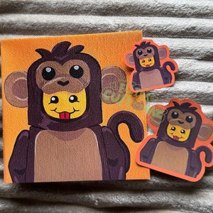 May include: A square canvas painting and two matching stickers featuring a yellow character wearing a dark brown monkey costume with a hood, large ears, and a tail. The character has a small red tongue sticking out against a bright orange background.