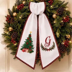 May include: A festive evergreen wreath decorated with pinecones, red berries, gold ornaments, and warm string lights. A white fabric ribbon with a red and green plaid border hangs from the center, featuring a printed Christmas tree and a letter G.