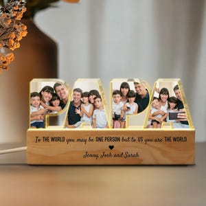 K&ouml;nnte beinhalten: Ein beleuchtetes Holzschild, das "PAPA" buchstabiert, mit einem Familienfoto in jedem Buchstaben. Das Schild tr&auml;gt die Botschaft "F&uuml;r die Welt bist du vielleicht nur eine Person, aber f&uuml;r uns bist du die Welt." und ist mit den Namen "Jenny, Josh und Sarah" personalisiert.