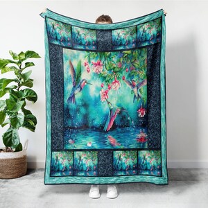 May include: A teal and blue hummingbird blanket featuring a vibrant watercolor design. The blanket showcases hummingbirds, pink flowers, and a reflective water scene. The border is a darker teal with a patterned design.