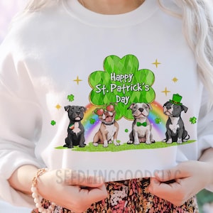 May include: White sweatshirt featuring four cartoon dogs celebrating St. Patrick's Day. The dogs are sitting on green grass with a rainbow and shamrock background. The phrase "Happy St. Patrick's Day" is written above the dogs.