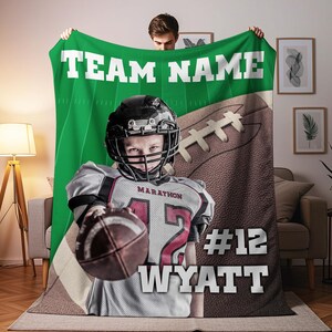 May include: A personalized football-themed blanket featuring a football player in a helmet and jersey holding a football. The blanket has a green background with the text "TEAM NAME" and the player's name and number.